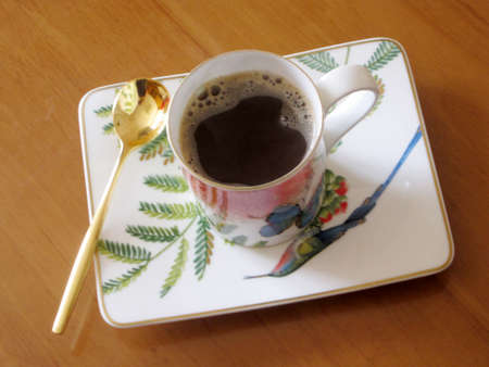 Beauty coffeecup standing on wooden table with dark tasty coffee. Brown old table consisting of liquid coffee in coffeecup, golden spoon, small plate. Porcelain coffeecup for coffee at antique table.の写真素材