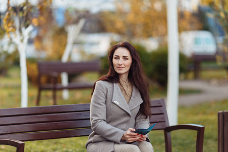 the girl sits on a bench and looks at the phone in autumn. behind blurred green park background. High quality photoの写真素材