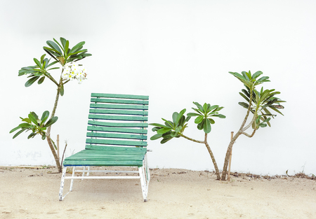 Old green wooden chaise longue against a white wall surrounded by plumeria treesの写真素材