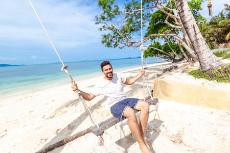 Young attractive happy mix raced man swinging on a swing on the shore of a bright tropical sea,...