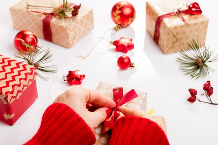 Process packing Christmas presents. White wooden table with red decorations, balls, candles, stars, female hands decorate gift red golden ribbon. New year, winter concept.の写真素材