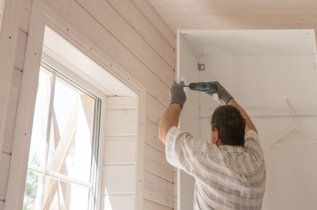 The process of assembling furniture, the master assembles a white cabinet using an electric drill in a room with a white wooden finish