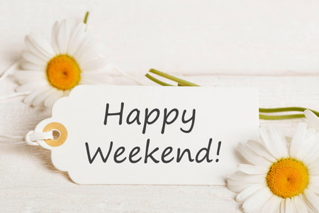 Happy Weekend message on white paper tag with daisies, minimal floral layout on light wood, clean summer concept for digital greeting cards, lifestyle blogs, printable templates, morning wishesの写真素材