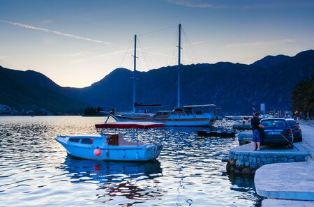 PERAST, MONTENEGRO - SEPTEMBER 13, 2017: Boats at waterfront of resort town of Perast against backdrop of mountains, evening view. Unknown people are on seafrontのeditorial素材