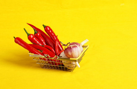 A shopping basket with vegetables and spices in the supermarket. Red pepper and fresh garlic in the shopping basket. Yellow background, with a place for inscription. High quality photoの写真素材