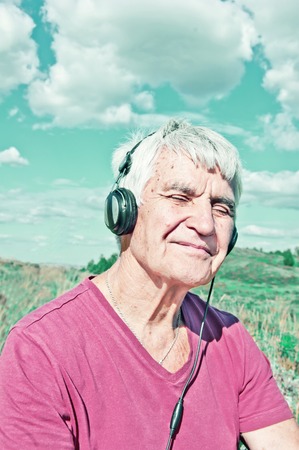 An elderly man with closed eyes in headphones listens to music Toned photo. Elderly smiling man sitting outdoor under cloudy sky. The influence of music on the brainの写真素材