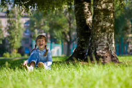 Laughing little girl sitting in grass near the treeの写真素材