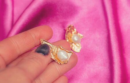 Hand holding Modern asymmetrical golden earrings with pearl beads over pink silk backgroundの写真素材