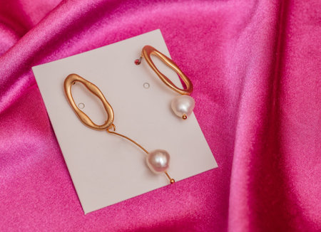 Modern asymmetrical golden earrings with pearl beads on pink silk backgroundの写真素材