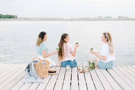three girls sitting on a wooden pier in front of the water. rest, picnic,lemonadeの写真素材