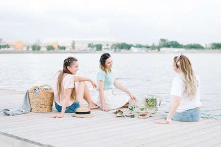 three girls sitting on a wooden pier in front of the water. rest, picnic,lemonadeの写真素材