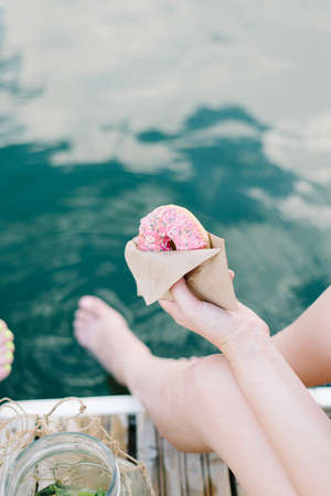 the girl holds in her hand an appetizing pink donut. outdoor recreation near the water. Dessert, pastry. craft packagingの写真素材
