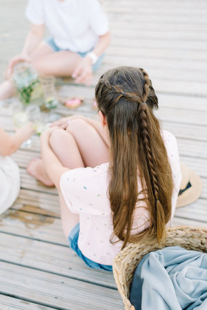 A woman in a pink t-shirt sitting in a wooden pier front of water, back view. Brown hair, pigtail...