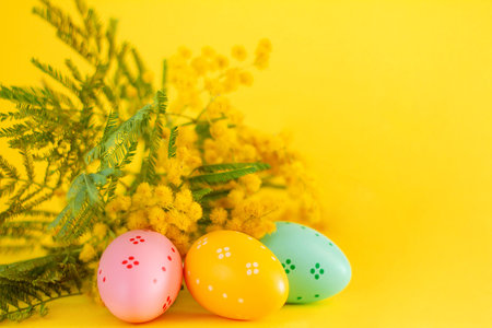 Colorful easter eggs and mimosa flowers on wooden table with copy space on yellow background. Easter