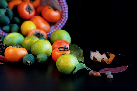 Fruits in basket: persimmons, lemons, pineapple guava with autumn leaves. Lemon Fresh fruit Persimmon Feijoa Fern Wicker basket Dark background Handsの写真素材