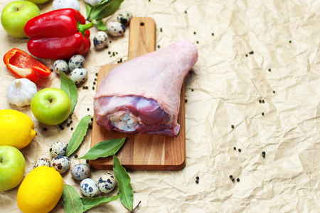 Raw turkey thigh garlic apples lemon red bell pepper quail eggs spices on a wooden board. Top view of a turkey, knife on a beige paper background. Culinary backgroundの写真素材