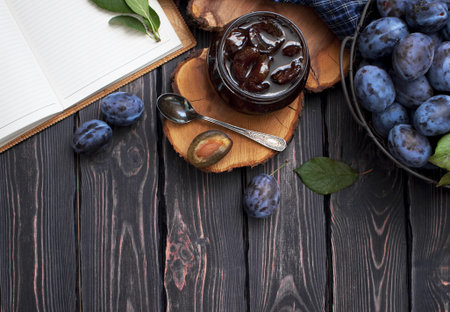 Homemade plum jam in a glass jar and fresh blue plums in a bowl on a dark rustic wooden background...