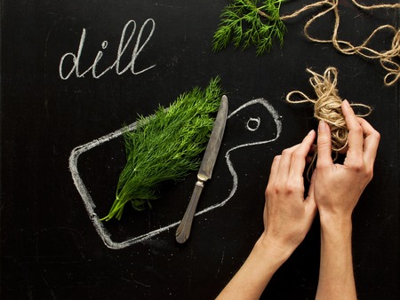 Concept of healthy eating, process of cooking, a healthy lifestyle. Hands of a woman, fresh dill on a hand-drawn cutting board, knife on a black background top view. Food backgroundの写真素材