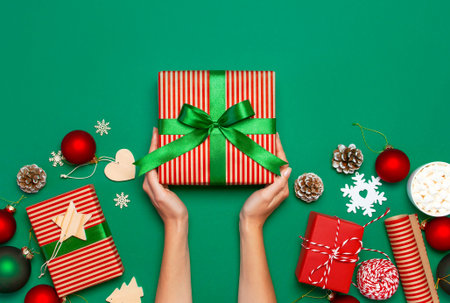 Woman hands holding present box, Christmas balls, toys, fir cones, ribbon on green background....