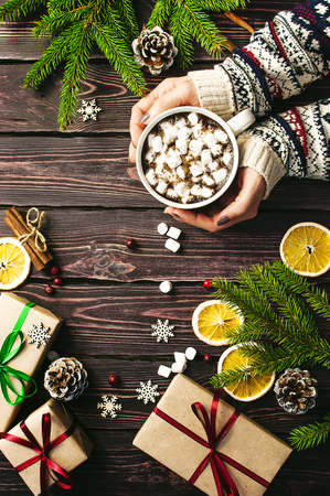 Female hands in woolen sweater with a cup of hot coffee with marshmallows, Christmas gifts, spruce branches, cones, orange slices, snowflakes, snow. Traditional festive decoration Christmas dark background top view. New Year Christmas composition.の写真素材