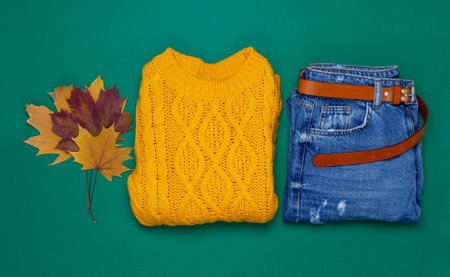 Female orange knitted sweater, blue jeans and autumn leaves on dark green background top view flat lay. Fashion Lady Clothes Set Trendy Cozy Knit Jumper Autumn accessories Female fashion lookの写真素材