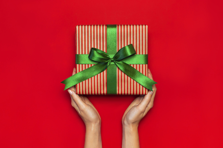 Gift box with green ribbon in female hands on red background top view flat lay. Holiday concept new year or Christmas gift box presents Xmas holiday Congratulations background with space for textの写真素材