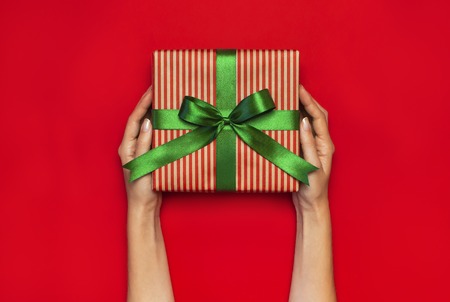 Gift box with green ribbon in female hands on red background top view flat lay. Holiday concept new year or Christmas gift box presents Xmas holiday Congratulations background with space for textの写真素材