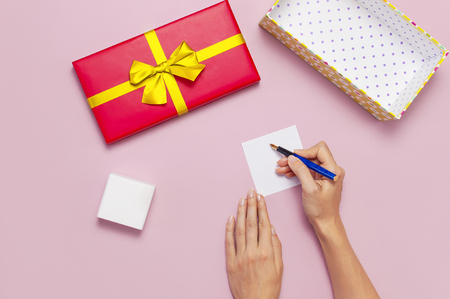 Flat lay top view womens hands write congratulations on empty sheet, red present box with gold ribbon on light pink background. Holiday concept birthday gift Congratulations background with copy spaceの写真素材