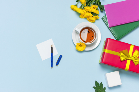 Red gift box with gold ribbon yellow chrysanthemums cup of tea notebook on blue background top view flat lay. Holiday concept, birthday gift, valentine day or anniversary Congratulations backgroundの写真素材