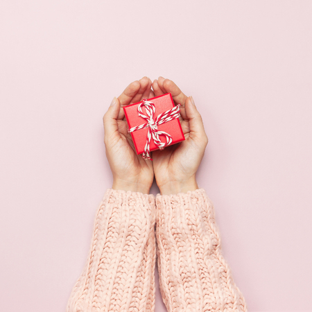 Female hands in pink knitted sweater holding a red gift box on pink background Flat lay style top view. Celebration, holiday, Birthday, Valentines day, Christmas, New Year copy spaceの写真素材