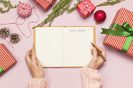 Female hands in pink knitted sweater are writing with pen in clean notebook plans for the new year, gift boxes fir branches on pink background flat lay. Christmas planning concept Holiday decorationsの写真素材