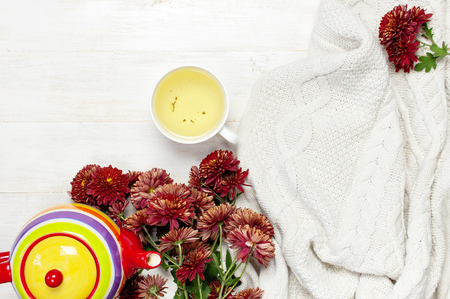 Cup of hot herbal tea, bright colored teapot, knitted plaid or sweater, red autumn chrysanthemum flowers on light wooden background top view Copy Space Cozy morning breakfast at homeの写真素材