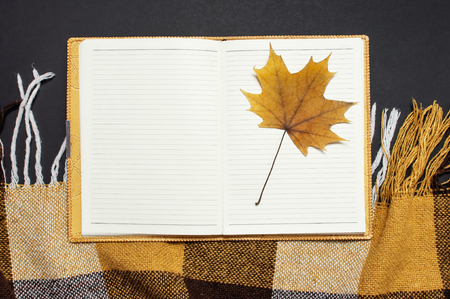 Opened notebook, yellow autumn maple leaf, orange brown checkered plaid on black background top view copy space. Autumn flat lay background. Autumn, fall halloween concept.の写真素材