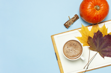 Opened notebook, orange pumpkin, pumpkin spiced latte or coffee in cup, cinnamon, yellow autumn maple leaf on blue background top view flat lay. Autumn or winter hot drink halloween concept.の写真素材