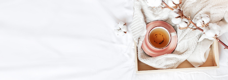 Cup of hot tea, wooden tray, branch of delicate cotton and knitted plaid sweater in bed. Cozy morning breakfast at home Lifestyle gentle female background Copy Space autumn winter conceptの写真素材