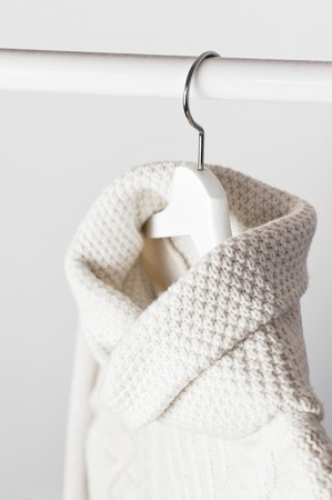 White knitted wool sweater on a white wooden hanger against the background of the light wall. Autumn and winter clothes. Store concept, sale design. Place for text.の写真素材