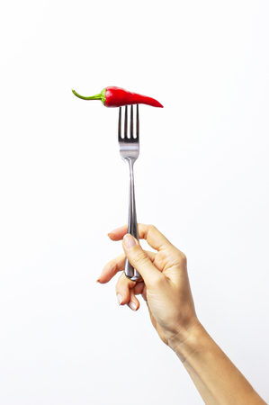 A female hand holds a fork with red chili pepper on white isolated background. Creative Food conceptの写真素材