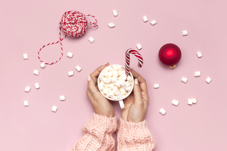 White mug with marshmallows in female hands in knitted sweater Candy Cane gifts boxes red ball on pink background top view Flat Lay Winter traditional food Festive decor celebration Christmas New Year.の写真素材