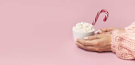 Female hands in knitted sweater holding cup of marshmallows and Christmas candy cane on pink background Flat Lay copy space Winter traditional food Festive decor celebration presents Xmas holiday.の写真素材