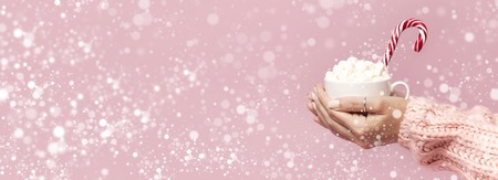 Female hands in knitted sweater holding cup of marshmallows and Christmas candy cane on pink background Flat Lay copy space Winter traditional food Festive decor celebration snowy effect.の写真素材