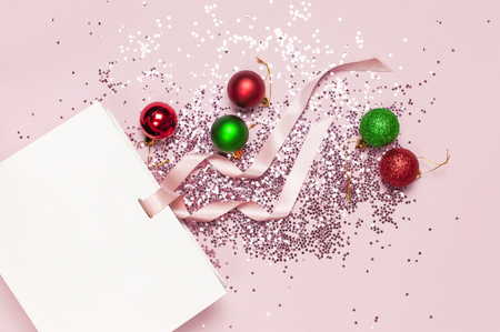 Flat lay top view White gift bag, holographic glitter confetti form of stars, red and green christmas balls on pink background. Greeting card Festive holiday decoration Congratulations New Yearの写真素材