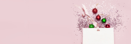 Flat lay top view White gift bag, holographic glitter confetti form of stars, red and green christmas balls on pink background. Greeting card Festive holiday decoration Congratulations New Yearの写真素材