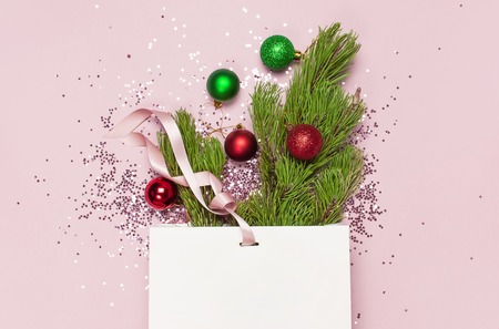 Flat lay top view White gift bag holographic glitter confetti red and green christmas balls green pine branches on pink background Greeting card Festive holiday decoration Congratulations NewYear.の写真素材