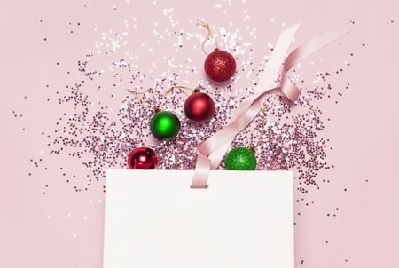 Flat lay top view White gift bag, holographic glitter confetti form of stars, red and green christmas balls on pink background. Greeting card Festive holiday decoration Congratulations New Yearの写真素材
