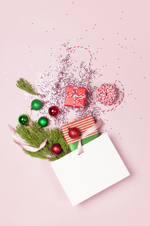 Flat lay top view White gift bag holographic glitter confetti red green christmas balls pine branches gift boxes on pink background. Greeting card Festive holiday decoration Congratulations New Yearの写真素材