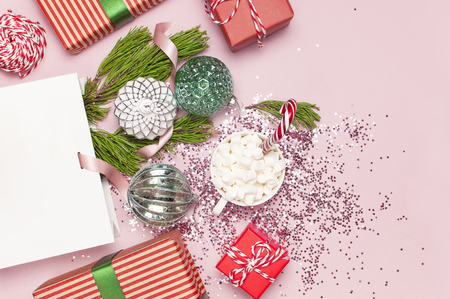Flat lay top view White gift bag holographic glitter confetti Vintage Christmas New Year balls pine branches gift boxes mug with marshmallows Candy Cane on pink background Greeting card decoration.の写真素材