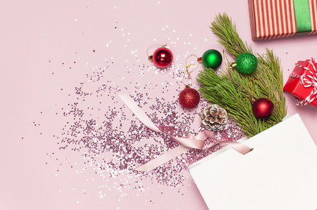 Flat lay top view White gift bag holographic glitter confetti red green christmas balls pine branches gift boxes on pink background. Greeting card Festive holiday decoration Congratulations New Yearの写真素材