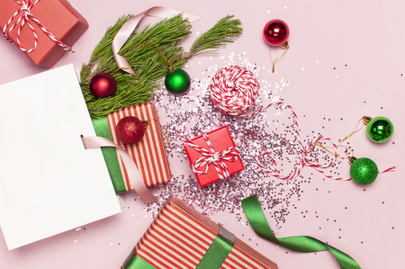 Flat lay top view White gift bag holographic glitter confetti red green christmas balls pine branches gift boxes on pink background Greeting card Festive holiday decoration.の写真素材