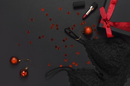 Black gift box with red ribbon, lace bra underwear, red lipstick, Christmas balls, holographic confetti on dark background top view flat lay. Female essential accessories gift for new year christmasの写真素材