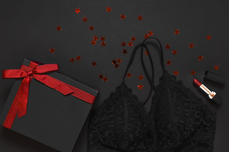 Black gift box with red ribbon, lace bra underwear, red lipstick, holographic confetti on dark background top view flat lay. Female essential erotic accessories, fashionable underwear gift to womanの写真素材